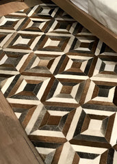 A cowhide patchwork rug with a geometric pattern featuring brown, grey, and white colors, laid out on a wooden floor.