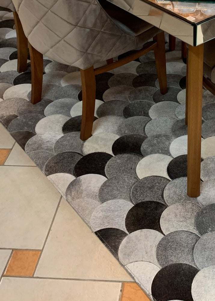 A cowhide patchwork rug with a geometric pattern in black, brown, and beige colors, laid out on a floor with a chair and a wooden table in the background.