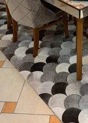 A cowhide patchwork rug with a geometric pattern in black, brown, and beige colors, laid out on a floor with a chair and a wooden table in the background.