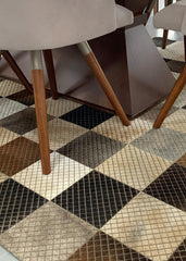A cowhide patchwork rug with a geometric block pattern in black, brown, beige, and white colors, laid out on a tiled floor with furniture legs visible.