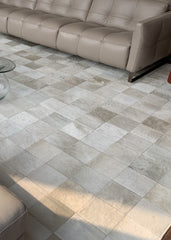 A cowhide patchwork rug in a champagne color, laid out on a tiled floor in a living room setting with a beige sofa and a glass coffee table in the background.