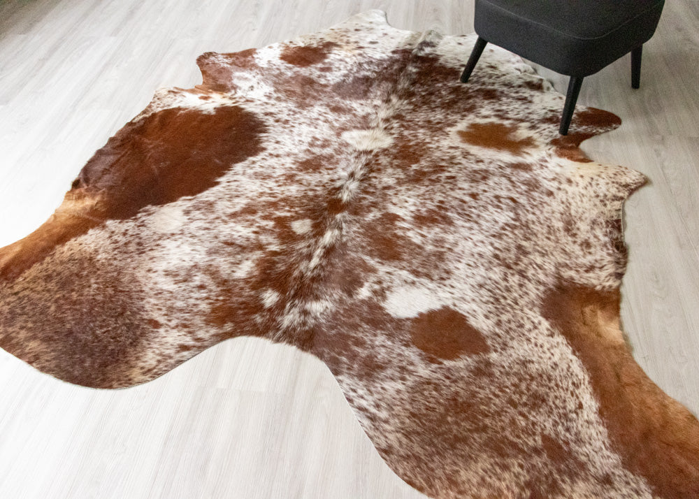 Brown and white cowhide rug on a light wooden floor with a chair in the corner.