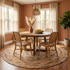 Dining room with wooden table and chairs on a colorful round jute rug, large windows, and a plant.