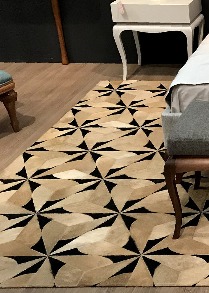 A cowhide patchwork rug with a geometric pattern in beige and black colors, placed on a wooden floor in a room with a white console table and a grey chair.