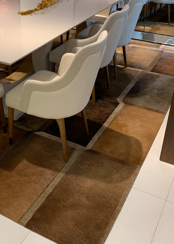 A brown cowhide patchwork rug laid out on a floor, with a white kitchen counter and chairs in the background.