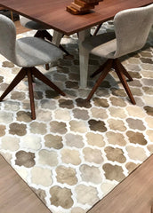 A cowhide patchwork rug with a geometric pattern in beige, champagne, and white colors, laid out on a wooden floor with a round wooden table and four chairs with upholstered seats in the background.