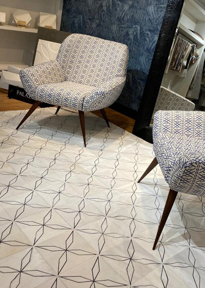 A cowhide patchwork rug with a geometric pattern in an off-white color, displayed in a showroom setting with a chair and mirror in the background.