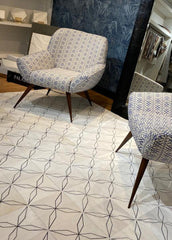 A cowhide patchwork rug with a geometric pattern in an off-white color, displayed in a showroom setting with a chair and mirror in the background.