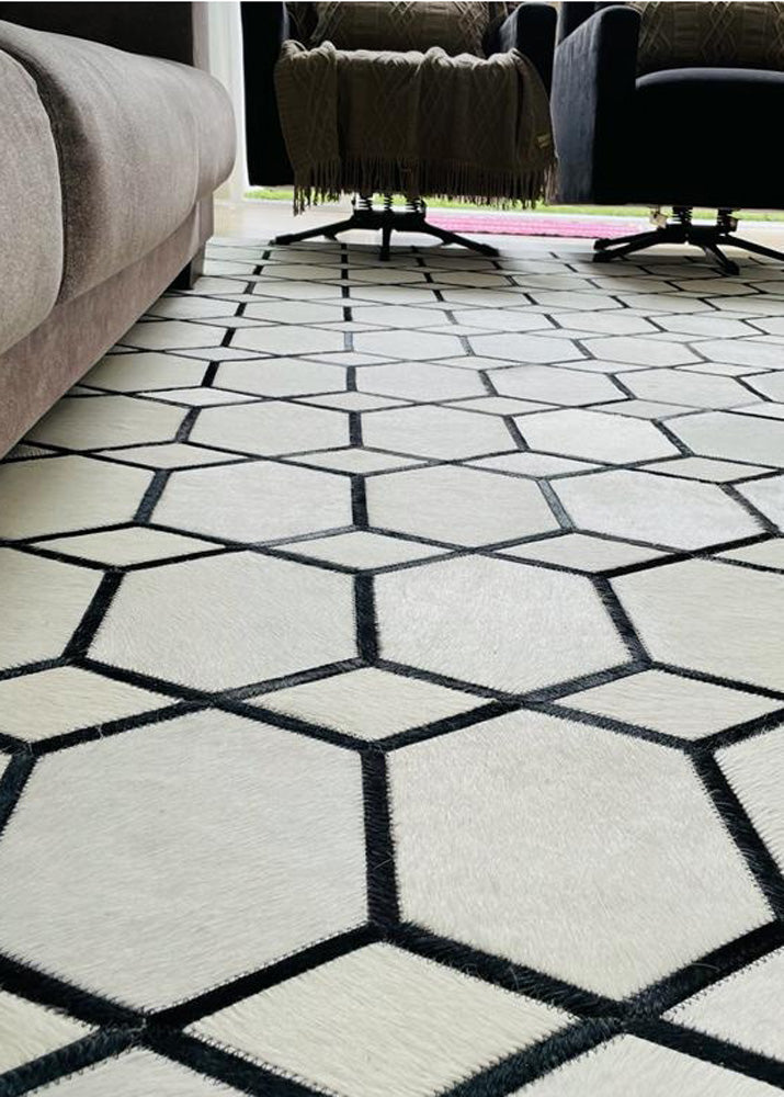 A cowhide patchwork rug with off white and black colors, laid out on a floor with a modern living room setting.
