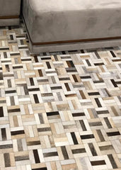 A cowhide patchwork rug featuring a geometric pattern in beige and brown colors, laid out on the floor with a couch in the background.