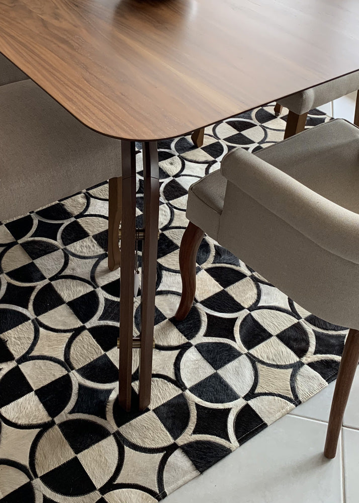 A cowhide patchwork rug with beige and black geometric patterns, placed on a wooden floor under a wooden table with a black and white patterned cushion.
