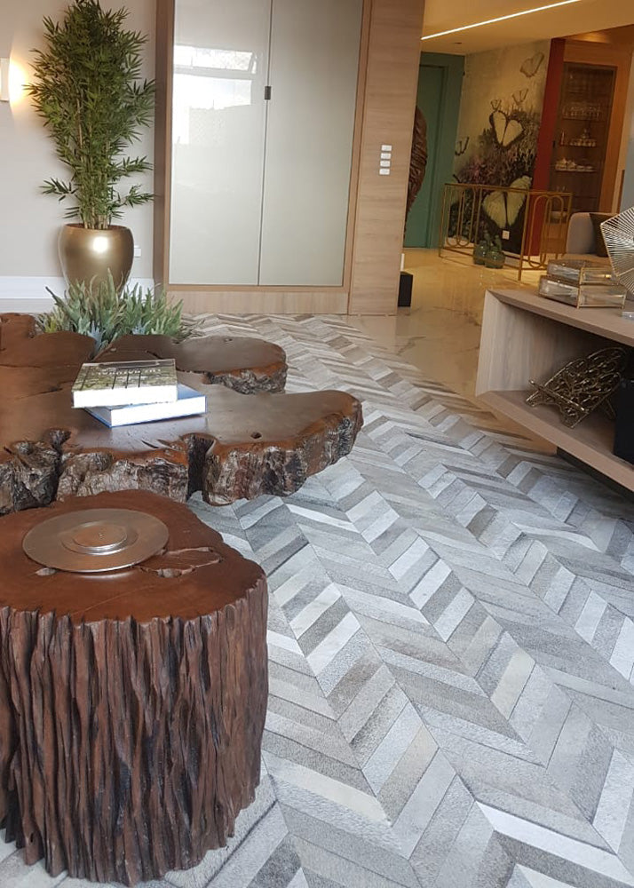 A cowhide patchwork rug featuring a geometric pattern in gray tones, laid out on a floor with wooden furniture and decorative plants.