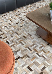 A cowhide patchwork rug featuring a geometric pattern in beige and brown colors, laid out on a floor with furniture in the background.