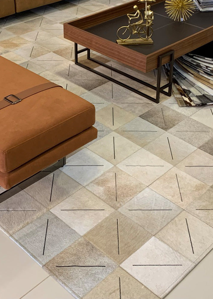 A cowhide patchwork rug in beige and off-white colors, laid out on a floor with a brown sofa and a wooden coffee table in the background.