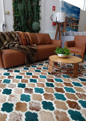 A multicolored patchwork cowhide rug with a geometric pattern, placed on a wooden floor in a living room setting with a brown sofa, brown accent chairs, and a wooden coffee table.